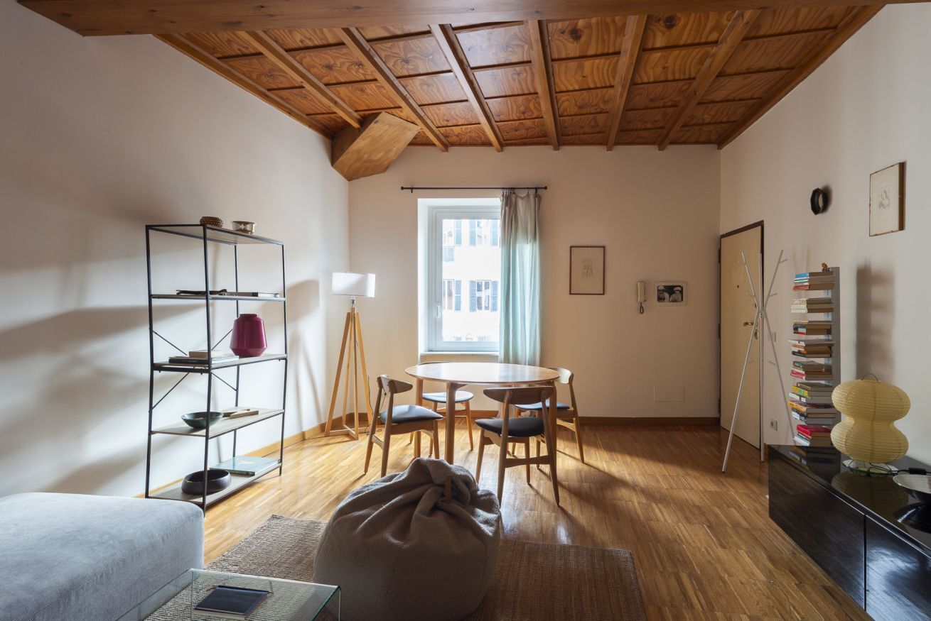 A room with a wooden ceiling, a round table with chairs, a bookshelf, and a window with curtains.