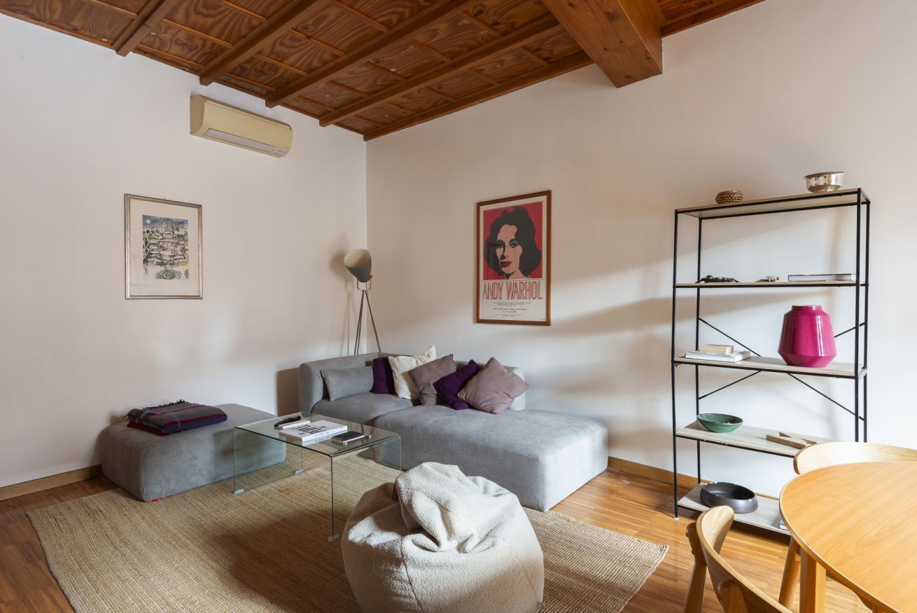 Una sala moderna con un sofá gris, una mesa de centro de vidrio, un sillón de peluche, una mesa de comedor de madera y una estantería metálica con objetos decorativos.