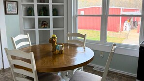 A dining area with a round wooden table, four white chairs, and a small dog on the table. There is a white shelving unit with various items on it, and a window with a view of a red barn and greenery outside. - 3-bedroom Bird's Nest Lodge in pleasant Cave Spring with AC and charm (Cave Spring)