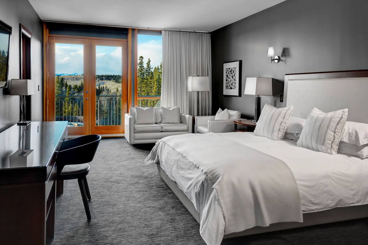A modern hotel room with a large bed, a desk, a chair, and a sofa. There is a balcony with a view of trees and mountains.