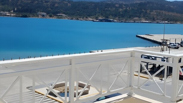 Vista costera con mar azul cristalino, un muelle blanco y montañas al fondo.
