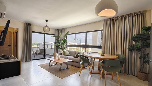 En moderne stue med sofa, sofabord, stole og spisebord. Der er et stort vindue med gardiner, en hængende lampe og en potteplante.