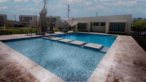 Et rektangulært swimmingpool med krystalklart blåt vand, omgivet af en fliseterrasse. Liggestole og parasoller findes i nærheden af poolen, og moderne bygninger ses i baggrunden.