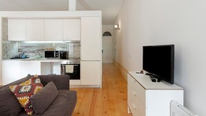 Un espace de vie compact comprenant un coin cuisine, un canapé agrémenté d’un plaid à motifs, un téléviseur et un meuble blanc.