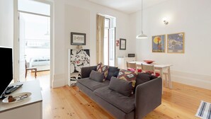 Un salon avec un canapé gris, un plancher en bois, un bureau blanc avec un ordinateur portable, une table blanche avec un bol rouge et un luminaire blanc au plafond.