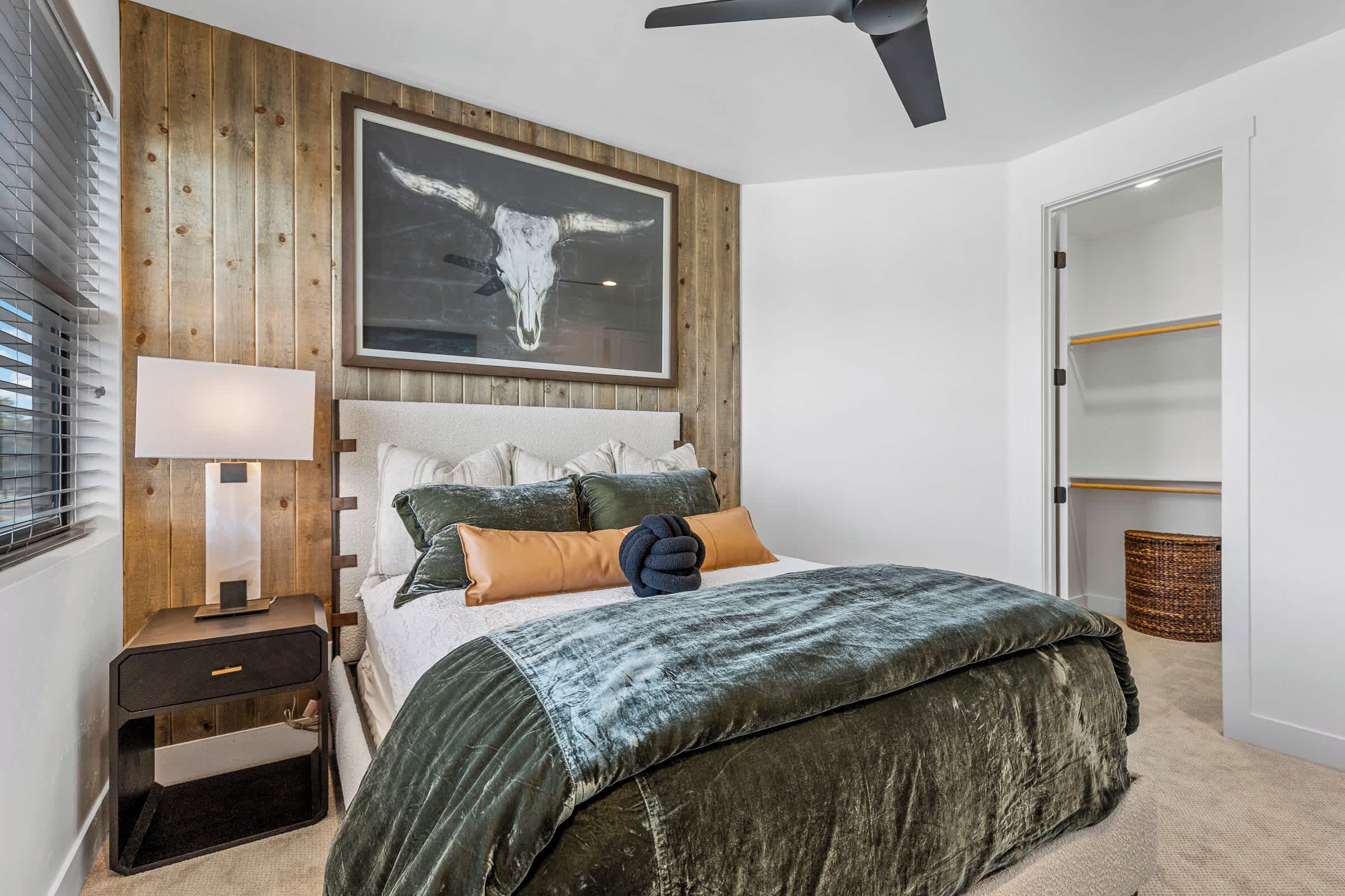 A bedroom with a wooden headboard, a large bull skull artwork, a bed with green and brown bedding, a nightstand with a lamp, and a ceiling fan.