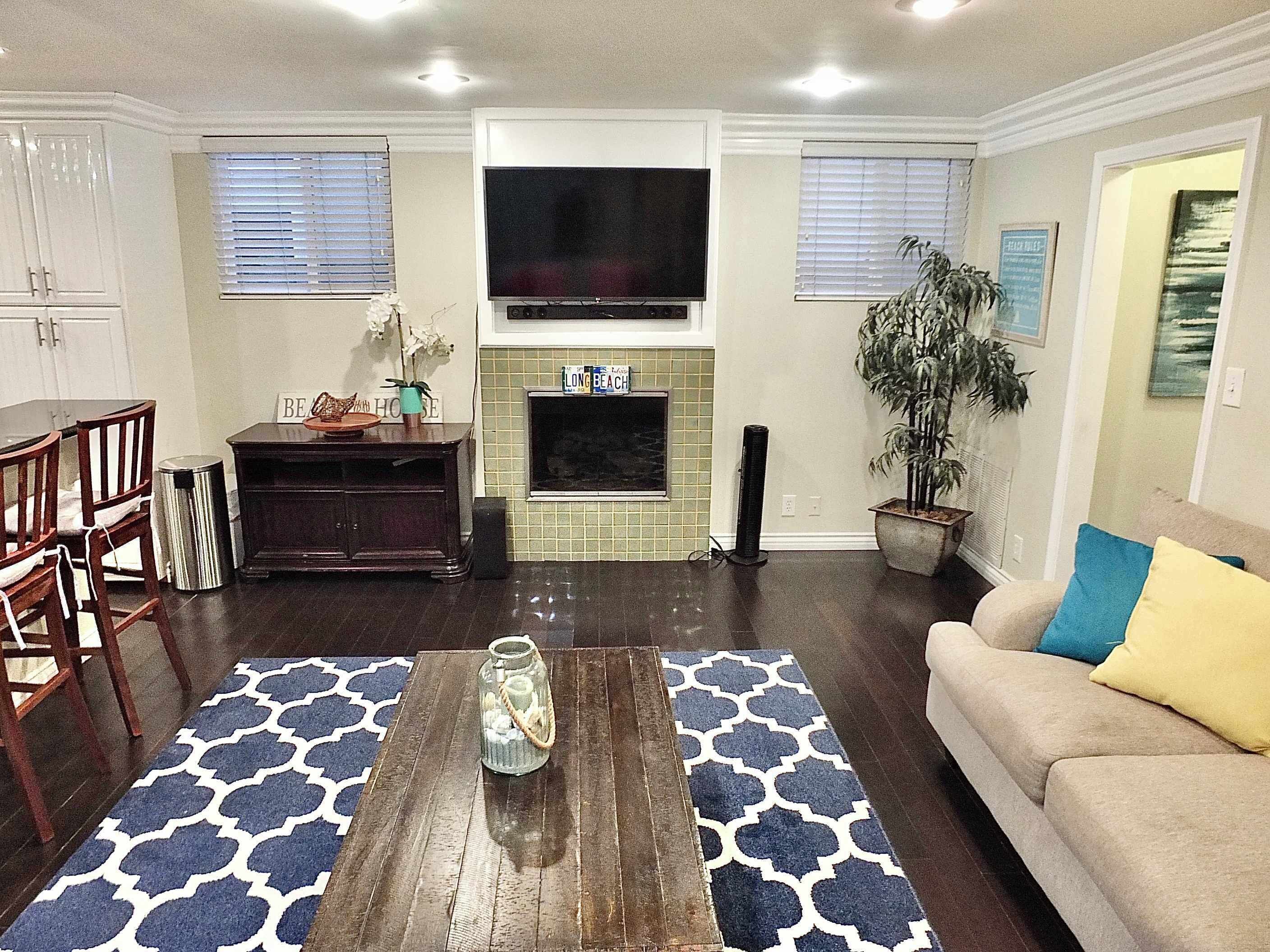 A living room with a fireplace, a TV mounted above it, a sofa with cushions, a coffee table, and a dining area with chairs.