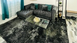 Un salon comprenant un canapé d’angle, une table basse en verre, un tapis noir, un tableau accroché au mur et une petite étagère ornée d’objets décoratifs.