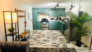 Une cuisine compacte avec des meubles de couleur turquoise, un four à micro-ondes, une cuisinière et un réfrigérateur. On y trouve un tapis à motifs, un banc et une plante en pot.