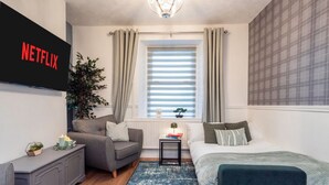 Et moderne soveværelse med en grå sofa, en seng med et blåt sengegavl, et lille bord med en plante og et vægmonteret TV, der viser 'NETFLIX'.
