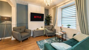 En moderne stue med to grå lænestole, en grøn sofa, et lille bord med bøger, en potteplante og et vægmonteret fjernsyn, der viser 'NETFLIX'.