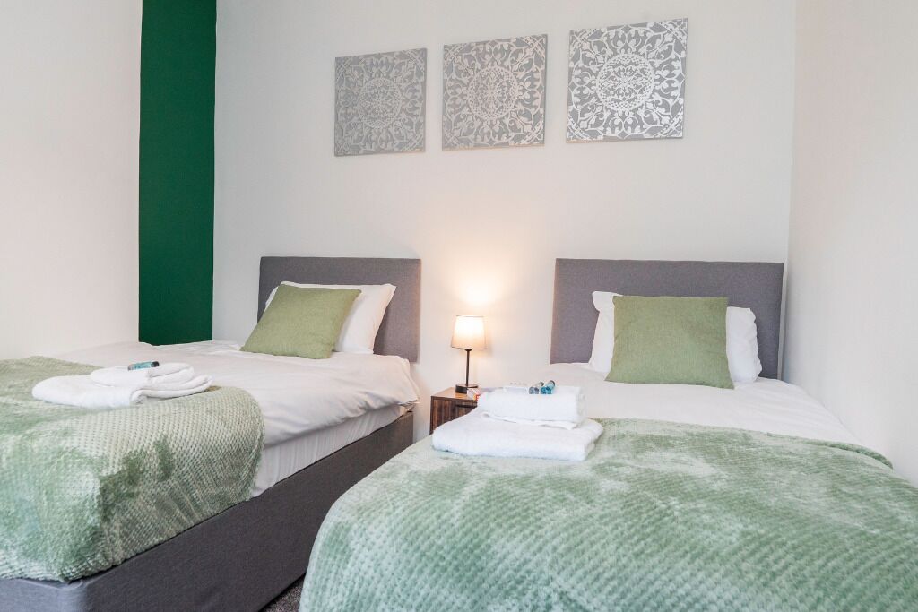 A hotel room with two beds, each with a green blanket and pillows, a nightstand with a lamp, and decorative wall art.
