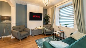 En moderne stue med to grå sofaer, en grøn sofa, et lille bord med bøger, en gulvlampe, en potteplante og et vægmonteret fjernsyn, der viser 'NETFLIX'.