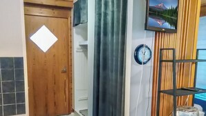 Una habitación con puerta de madera, alfombra a cuadros blanco y negro, cortina oscura, reloj en la pared y estante con un control remoto.