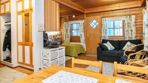 Ein gemütliches Blockhäuschen im Inneren mit Holzwänden und -decke, einem Fernseher auf einem weißen Schrank, einem grünen Bett, einem dunklen Sofa und einem Korbstuhl.