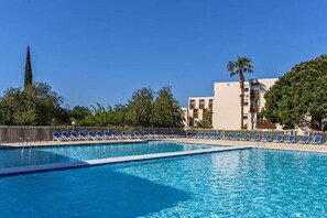 Ein Swimmingpool mit blauem Wasser, umgeben von Liegestühlen und einem Gebäude mit mehreren Etagen und Balkonen.