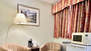 Una stanza piccola con due poltrone beige, un tavolo in legno, una lampada, una macchina per il caffè, un forno a microonde e un tappeto decorato.