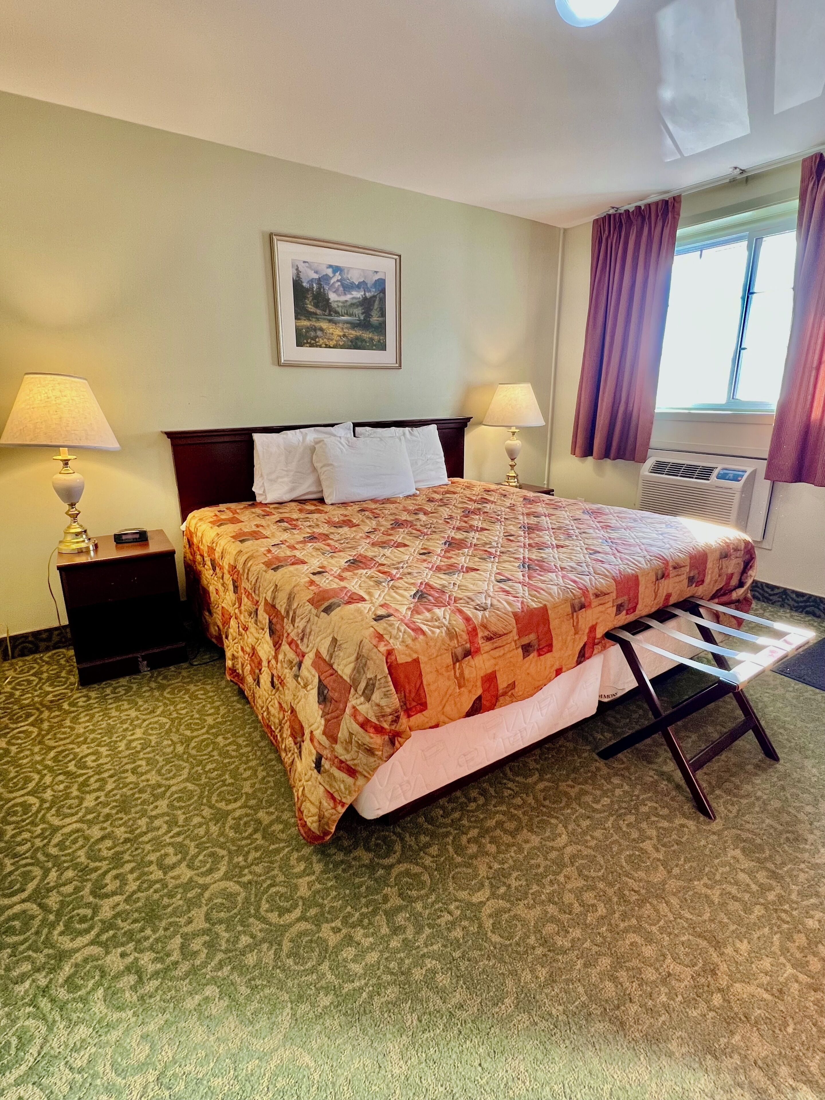 A hotel room with a bed, two bedside lamps, a window with curtains, a wall-mounted picture, and a carpeted floor.