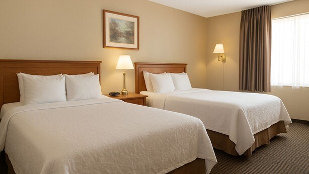 A hotel room with two beds, a nightstand with a lamp, a painting on the wall, and a window with curtains. - Rocky Mountain Lodge - 2 Queen Bed Size (Renfrew)