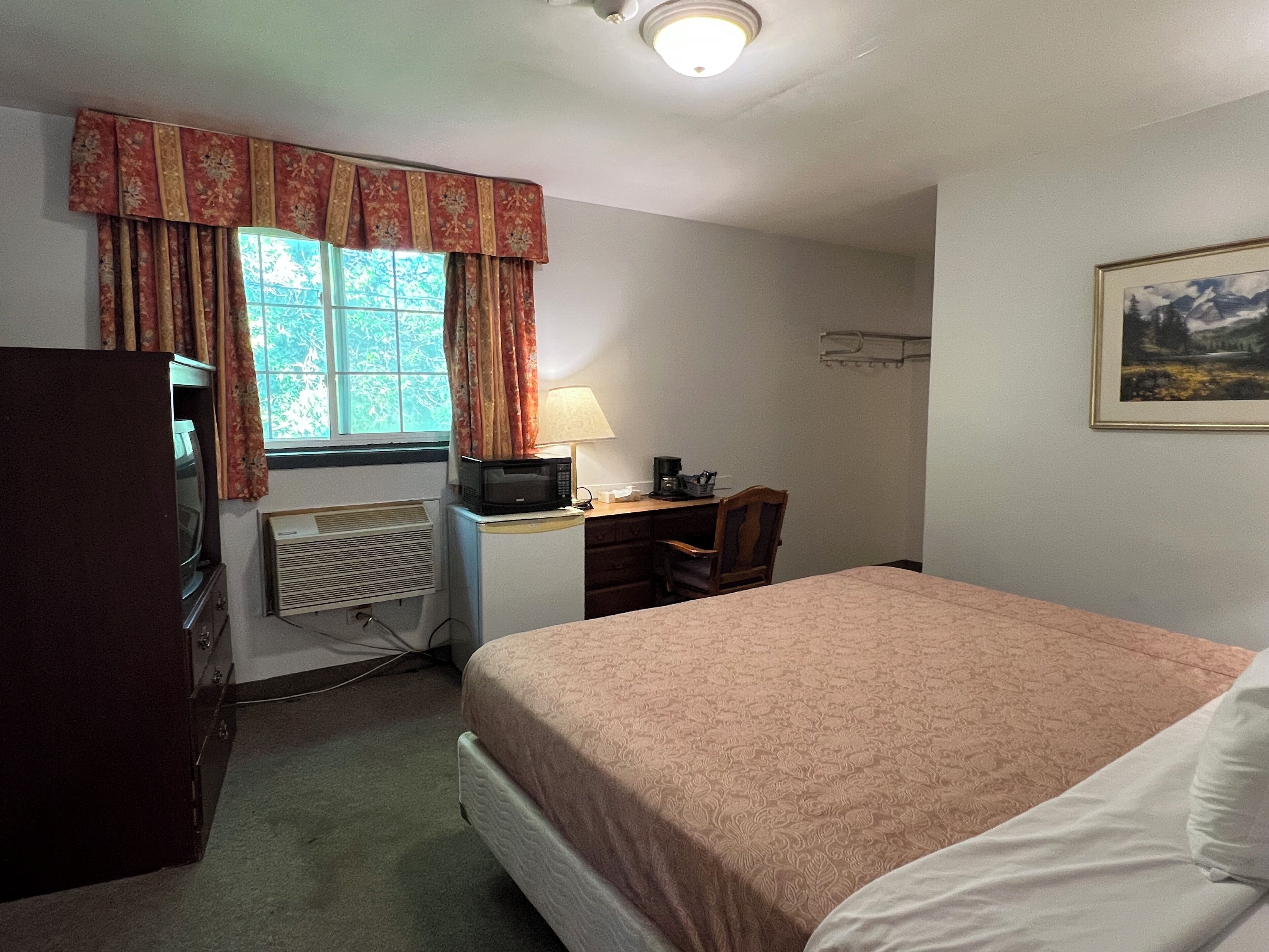 A compact hotel room with a bed, a desk with a computer, a chair, a television, and a window with curtains.