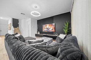 A modern living room with a large flat-screen TV, dark grey sofas, a patterned ottoman, and a minimalist staircase. - 8BR house sleeps 24 with spa and private pool (Kissimmee)