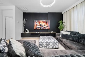 A modern living room with a flat-screen TV, dark sofas, and a patterned ottoman. - 8BR house sleeps 24 with spa and private pool (Kissimmee)