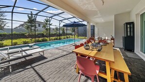 Uma área externa coberta com piscina, mesa de madeira com cadeiras e vista para uma casa ao fundo.