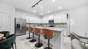 Cozinha moderna com armários brancos, geladeira de aço inoxidável, micro-ondas e um espaço bar com banquetas.