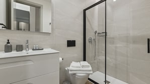 Un baño moderno con ducha de vidrio, un lavamanos blanco con espejo encima y un inodoro con una pila de toallas.