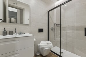 Ein modernes Badezimmer mit einer gläsernen Duschkabine, einem weißen Waschbecken mit Spiegel darüber und einer Toilette mit Handtüchentürmchen.