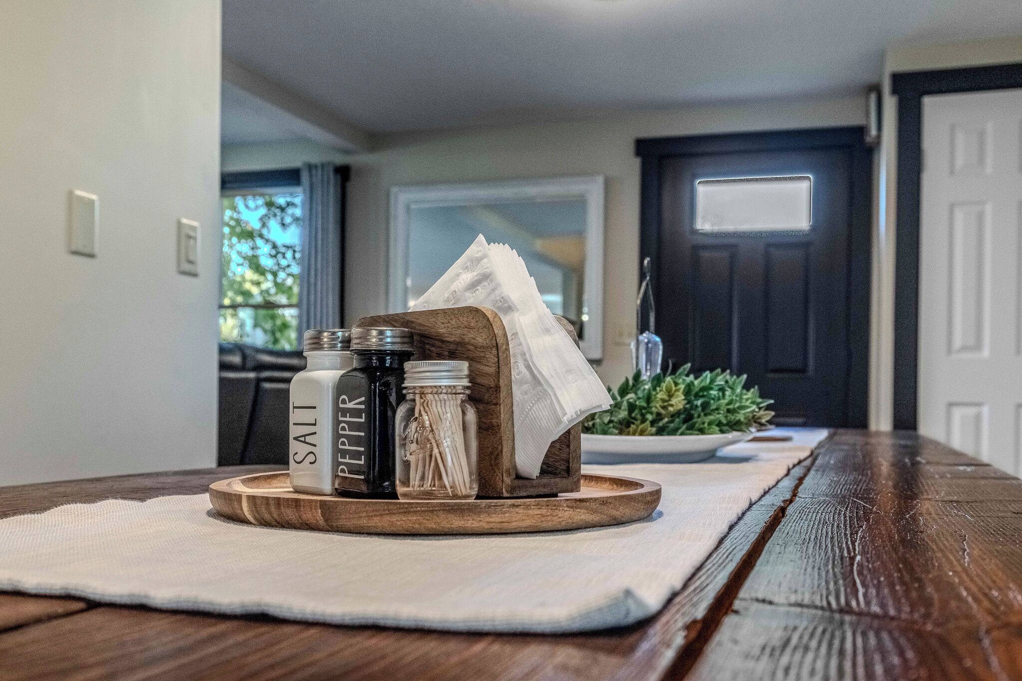 Una mesa de madera con mantel blanco, saleros y pimenteros, un tarro de cristal y un posavasos de madera con una planta.