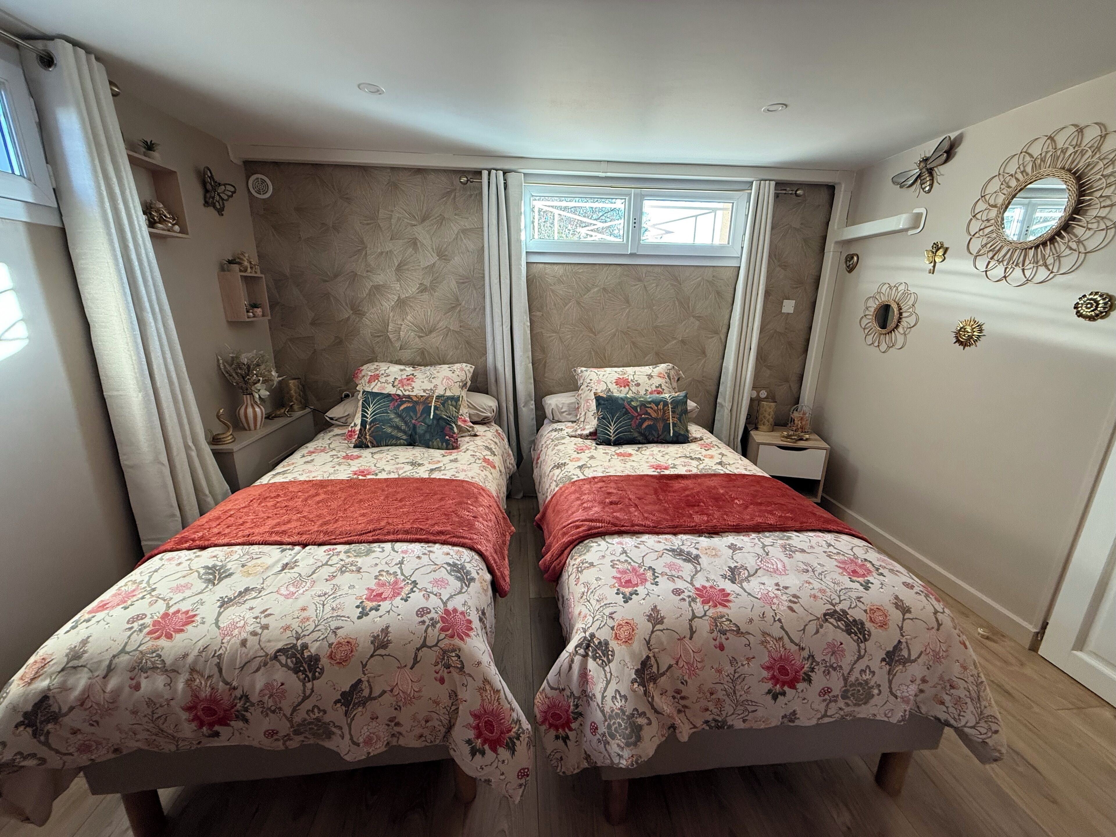 Une chambre avec deux lits, chacun agrémenté de draps à motifs floraux et recouvert d’un plaid rouge. On y trouve des tables de chevet ornées d’objets décoratifs et une fenêtre avec des rideaux.