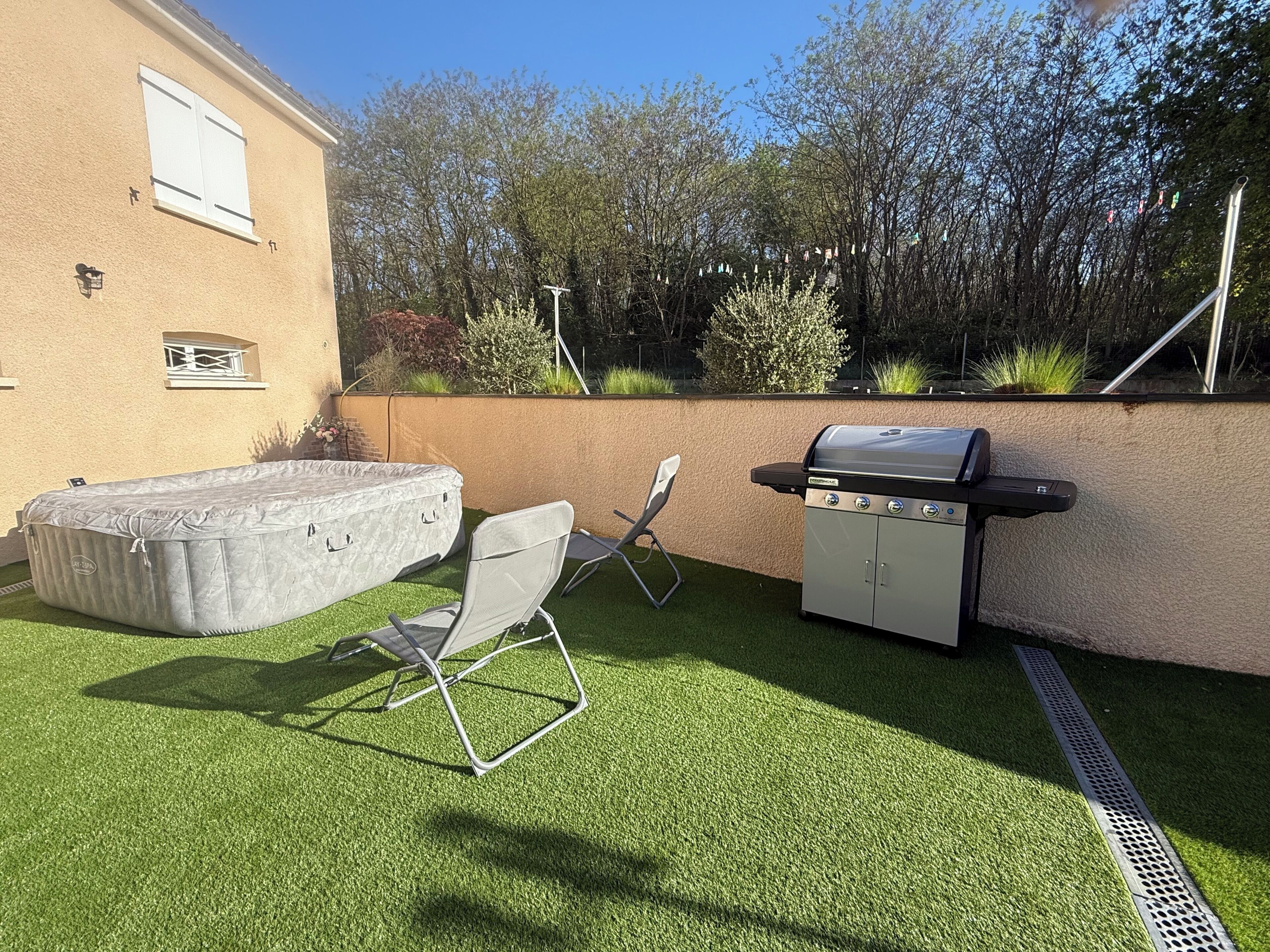 A backyard with artificial grass, a barbecue grill, a folding chair, and a hot tub.
