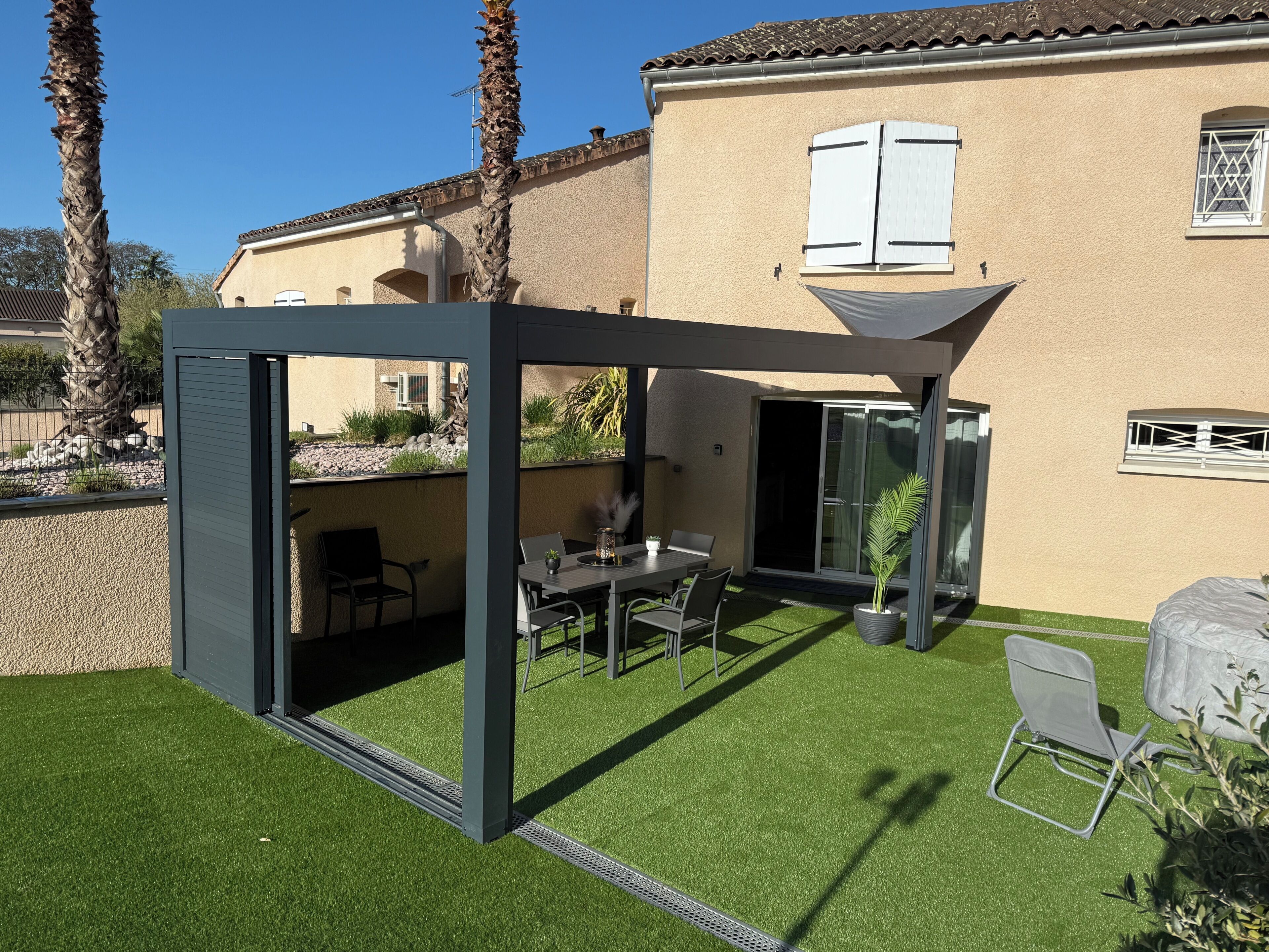 A modern outdoor patio with a glass enclosure, a dining table, chairs, and a small potted plant.