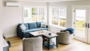A modern living room with a blue sofa, two armchairs, a coffee table, and a bookshelf. Large windows provide natural light and a view of the outdoors. - Luxury Roxbury Pond Lakefront (Roxbury)