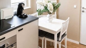 Una cuina compacta amb una cafetera, una taula blanca i caires, i una finestra amb persianes.