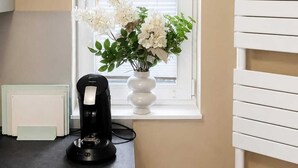 Una cuina amb una finestra, un radiador blanc, una cafetera negra i un vas de flors blanc.