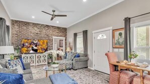 A living room with a brick wall, a television, a ceiling fan, and a dining area with a table and chairs. - Southern Sisters Retreat - The Lisa: Stylish, Cozy, Near MSU, Fire Pit + Grill (Starkville)
