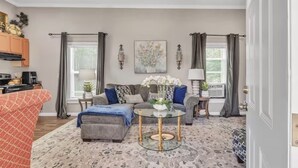 A living room with a sofa, ottoman, coffee table, and artwork on the wall. - Southern Sisters Retreat - The Lisa: Stylish, Cozy, Near MSU, Fire Pit + Grill (Starkville)