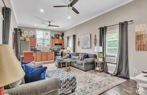 A spacious living room with a grey sofa, patterned ottoman, and a coffee table. There is a kitchen in the background with wooden cabinets and a dining area. - Southern Sisters Retreat - The Lisa: Stylish, Cozy, Near MSU, Fire Pit + Grill (Starkville)