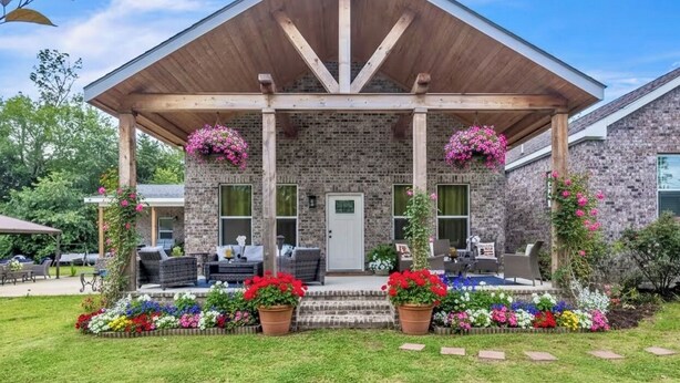 A brick house with a wooden porch, outdoor seating, and colorful flower beds. - Southern Sisters Retreat - The Lisa: Stylish, Cozy, Near MSU, Fire Pit + Grill (Starkville)
