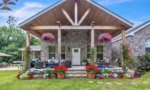 A brick house with a wooden porch, outdoor seating, and colorful flower beds. - Southern Sisters Retreat - The Lisa: Stylish, Cozy, Near MSU, Fire Pit + Grill (Starkville)
