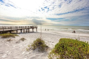 Ein Strand mit einer hölzernen Pier, die sich ins Meer erstreckt, feinem Sand und vereinzelter Vegetation.