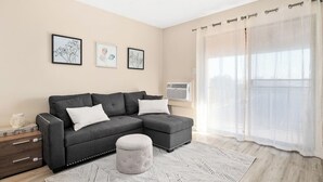 Una sala de estar moderna con un sofá seccional gris, un reposapiés blanco y una alfombra estampada. Hay tres obras de arte enmarcadas en la pared, y un ventanal con cortinas traslúcidas.
