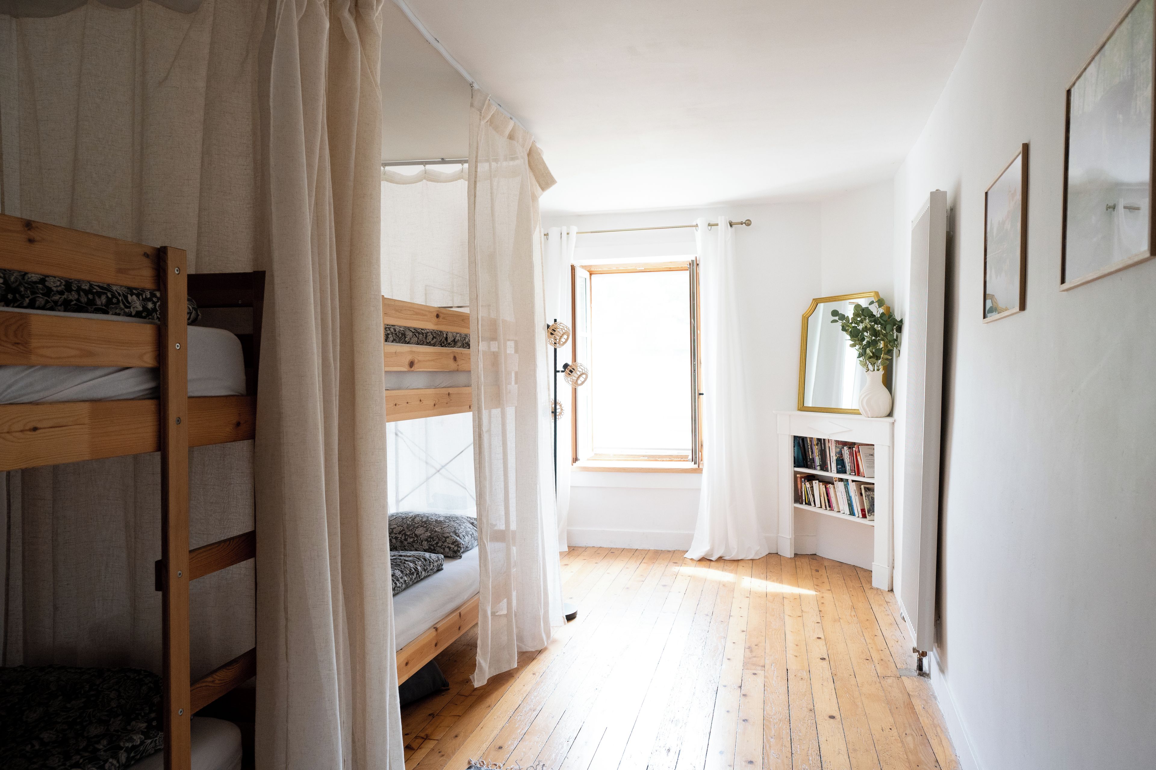 A room with bunk beds, a window with curtains, a bookshelf, and a mirror on the wall.
