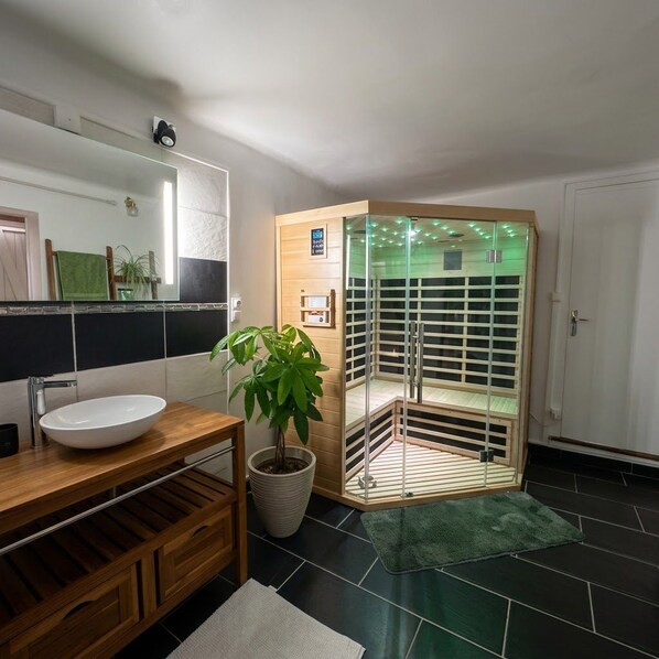 A bathroom with a wooden vanity, a white sink, a potted plant, a glass-enclosed shower, and a green mat on the floor.