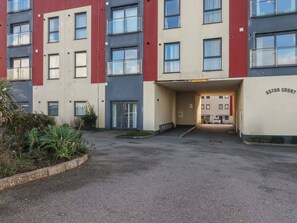 A multi-story building with a flat facade, a parking area in front, and a sign indicating a court. - Sea Snug (Newquay)