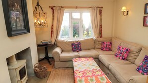 Un salon chaleureux avec un canapé beige, des coussins colorés, un plaid au point de crochet, une petite table avec des livres, un panier en osier et un poêle ancien.