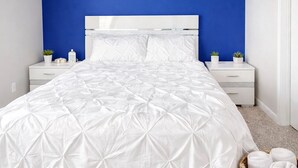 A bedroom with a white bed, a blue accent wall, and a small bedside table with a plant. - 5-STAR Sarasota Stay Luxur 2BR/2BA (Sarasota)