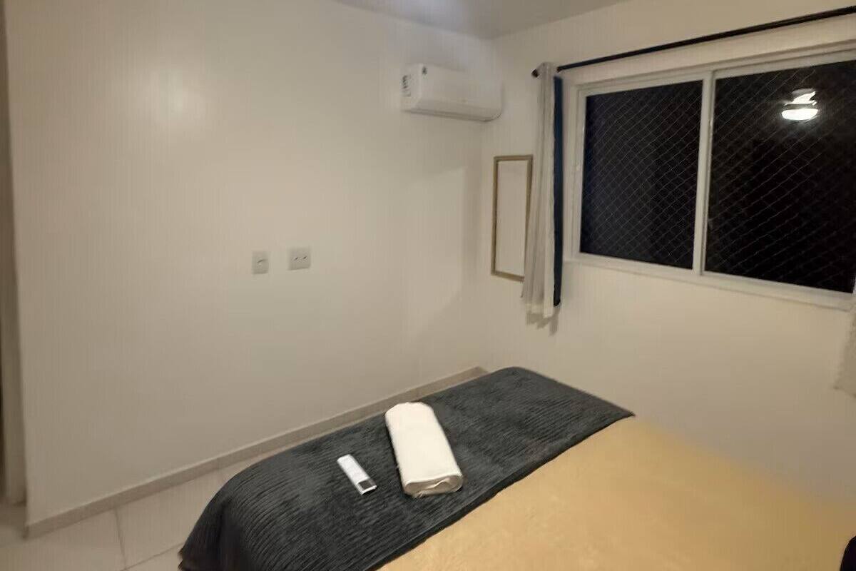 Een slaapkamer met een bed, een plafondventilator, een airconditioning, een raam met zonwering en een nachtkastje met een lamp en een opgerolde handdoek.